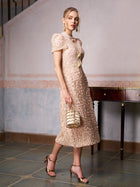 Woman in the Social Queen Floral Embroidered Lace Dress with short sleeves, accessorized with a textured handbag, gold heels, and earrings by a wooden table—an elegant choice for wedding guests.