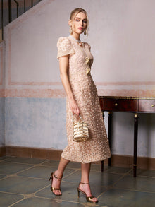 Woman in the Social Queen Floral Embroidered Lace Dress with short sleeves, accessorized with a textured handbag, gold heels, and earrings by a wooden table—an elegant choice for wedding guests.