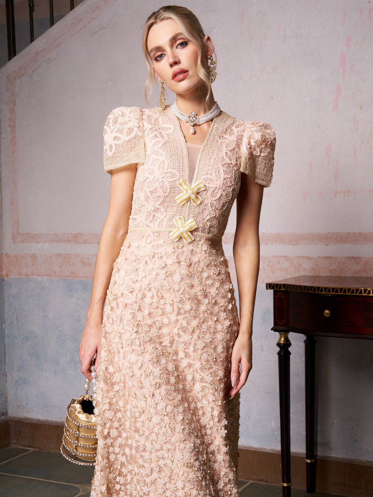 A woman indoors holds a beaded handbag while wearing the Social Queen Floral Embroidered Lace Dress, featuring light pink lace, layers, puffed sleeves, and floral embroidery.