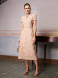 A woman poses indoors near a wooden table in the Social Queen Floral Embroidered Lace Dress—a light pink, knee-length lace dress with short puff sleeves—styled with sandals and a small handbag. Ideal as a wedding guest dress.