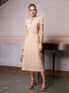 A woman poses indoors near a wooden table in the Social Queen Floral Embroidered Lace Dress—a light pink, knee-length lace dress with short puff sleeves—styled with sandals and a small handbag. Ideal as a wedding guest dress.