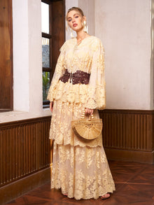 A woman poses indoors near wooden-framed windows, holding a wooden handbag. She wears Social Queen's Floral Embroidered Lace Layered Skirt: a long, beige skirt with 3D floral embroidery, paired with a wide dark belt.