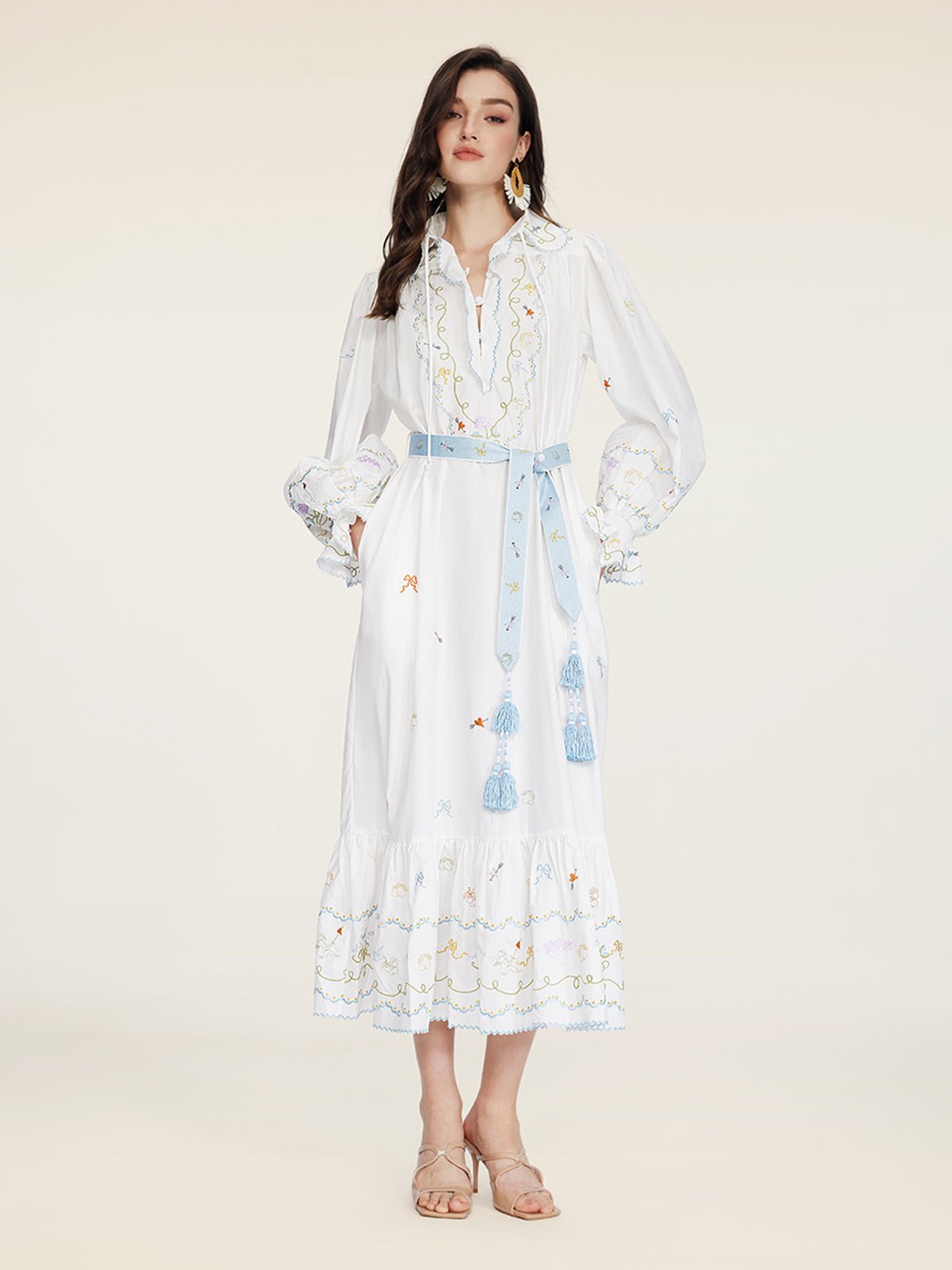 A woman wears the Social Queen Floral Embroidered Lantern Sleeve Dress with a blue tassel belt and beige sandals, standing against a plain light background—an ideal choice for summer sophistication.