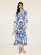 A woman stands against a plain background in the Social Queen Floral Embroidered Lantern Sleeve Dress, featuring white and blue floral patterns, a deep V-neck, and long sleeves. She holds a small silver handbag and wears silver sandals with jewelry.