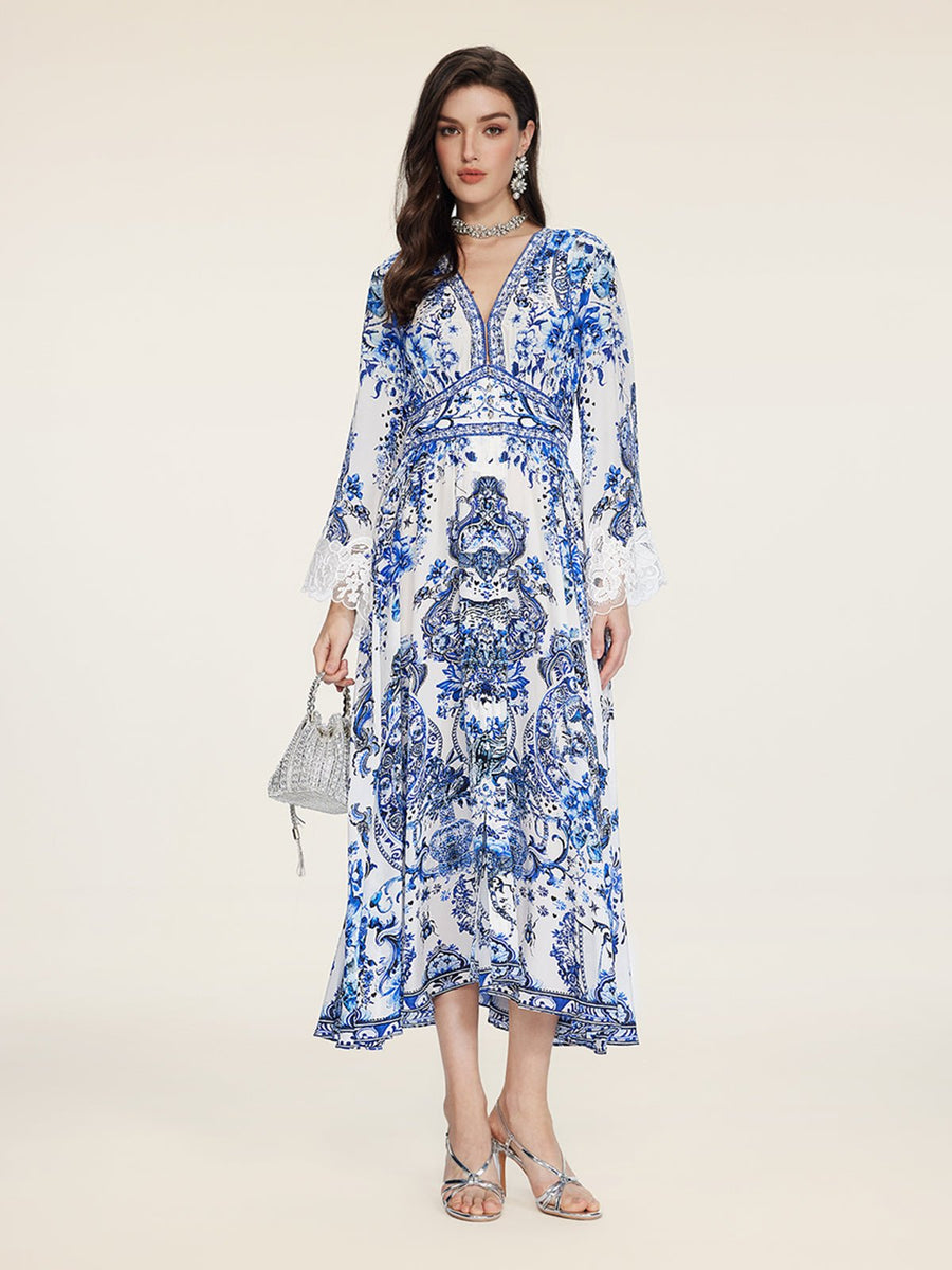 A woman stands against a plain background in the Social Queen Floral Embroidered Lantern Sleeve Dress, featuring white and blue floral patterns, a deep V-neck, and long sleeves. She holds a small silver handbag and wears silver sandals with jewelry.