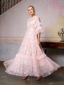A woman stands indoors near a staircase and table with a candelabra, wearing the Social Queen Floral Embroidered Layered Tulle Dress in light pink with ruffles and floral embellishments.