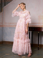 A woman poses indoors, touching her head, in the Social Queen Floral Embroidered Layered Tulle Dress—a long, light pink gown with ruffled sleeves and hem—perfect as a wedding guest dress.