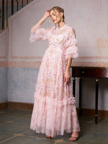 A woman poses indoors, touching her head, in the Social Queen Floral Embroidered Layered Tulle Dress—a long, light pink gown with ruffled sleeves and hem—perfect as a wedding guest dress.
