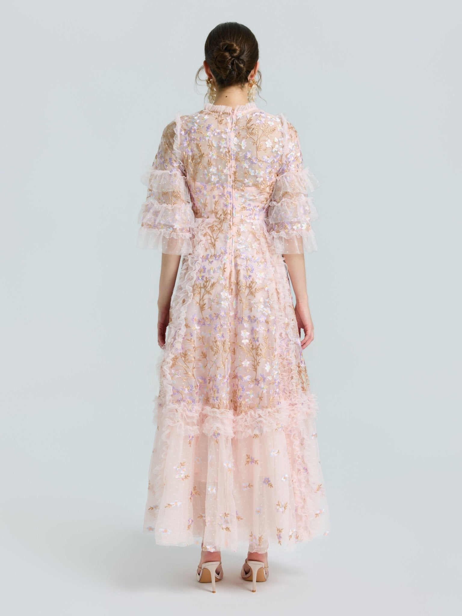 A woman faces away, wearing the Social Queen Floral Embroidered Layered Tulle Dress with ruffled sleeves and hem, nude heels, and a sheer pink floral design against a plain light background.