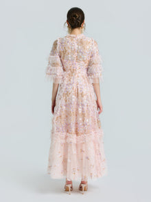 A woman faces away, wearing the Social Queen Floral Embroidered Layered Tulle Dress with ruffled sleeves and hem, nude heels, and a sheer pink floral design against a plain light background.