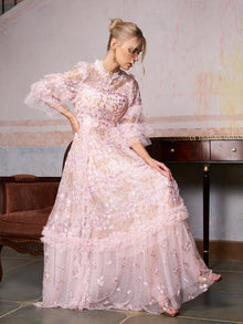 A woman in the Social Queen Floral Embroidered Layered Tulle Dress sits on a brown vintage-style couch with her hand on the skirt, next to a wooden table and against a textured wall.