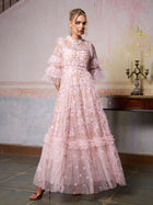 A woman in Social Queen's Floral Embroidered Layered Tulle Dress with ruffled sleeves stands indoors by a wooden table with a candelabra—an elegant choice for a wedding guest.