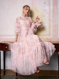 A woman in the Social Queen Floral Embroidered Layered Tulle Dress sits on a table, holding a small floral handbag and wearing strappy sandals, against a textured wall background.
