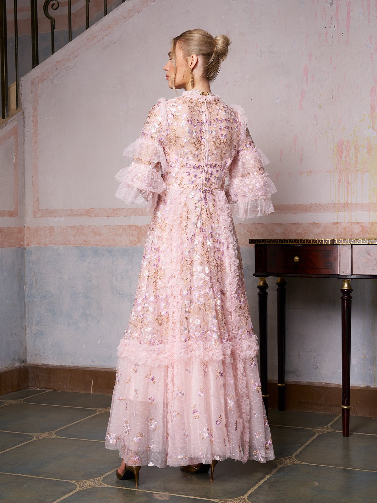 A woman stands indoors by a staircase and table, wearing the Social Queen Floral Embroidered Layered Tulle Dress—an elegant choice with sheer pink fabric, floral embroidery, and ruffled sleeves for wedding guests.