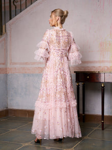 A woman stands indoors by a staircase and table, wearing the Social Queen Floral Embroidered Layered Tulle Dress—an elegant choice with sheer pink fabric, floral embroidery, and ruffled sleeves for wedding guests.