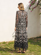A woman stands on grass facing a white wall, wearing the Social Queen Floral Embroidered Mesh Dress with sheer panels and sandals—a chic choice for a wedding guest.