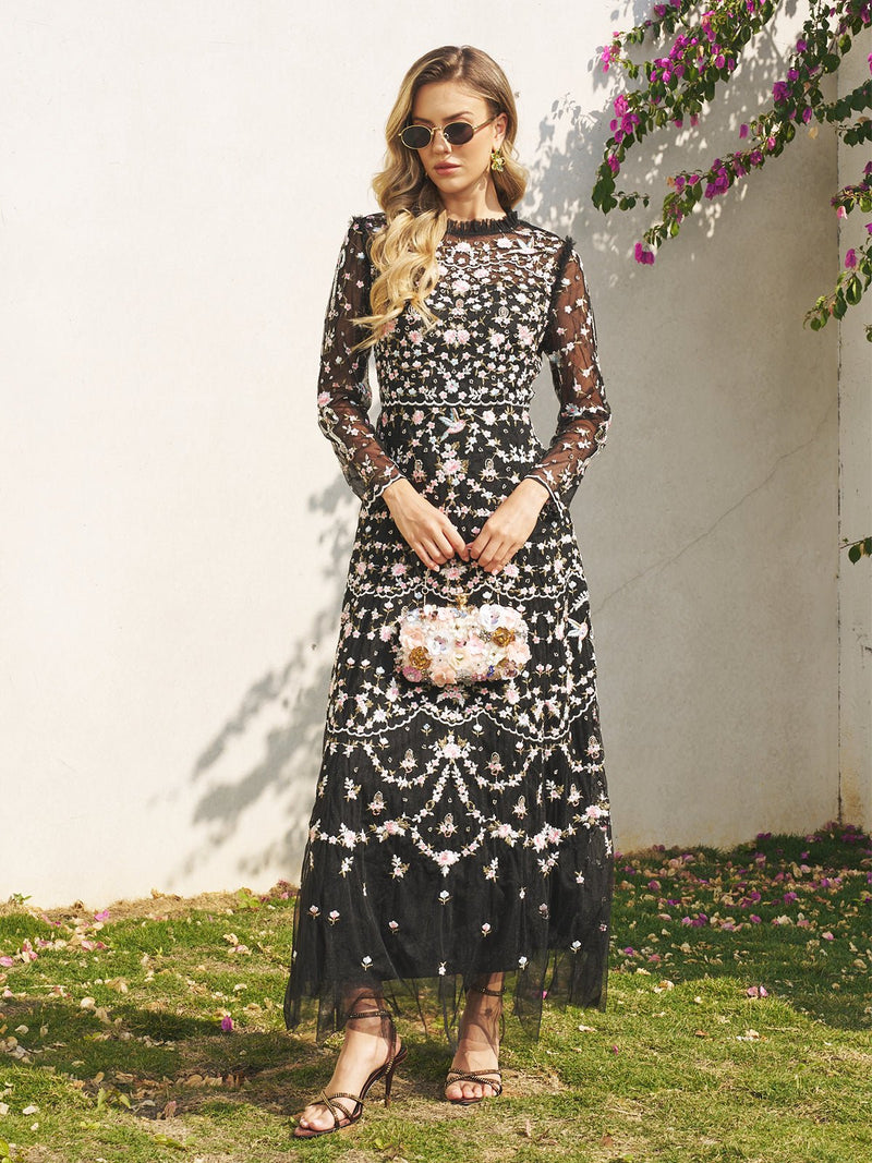 A woman in sunglasses wears the Social Queen Floral Embroidered Mesh Dress and holds a small handbag, standing on grass near a white wall with flowers and greenery.