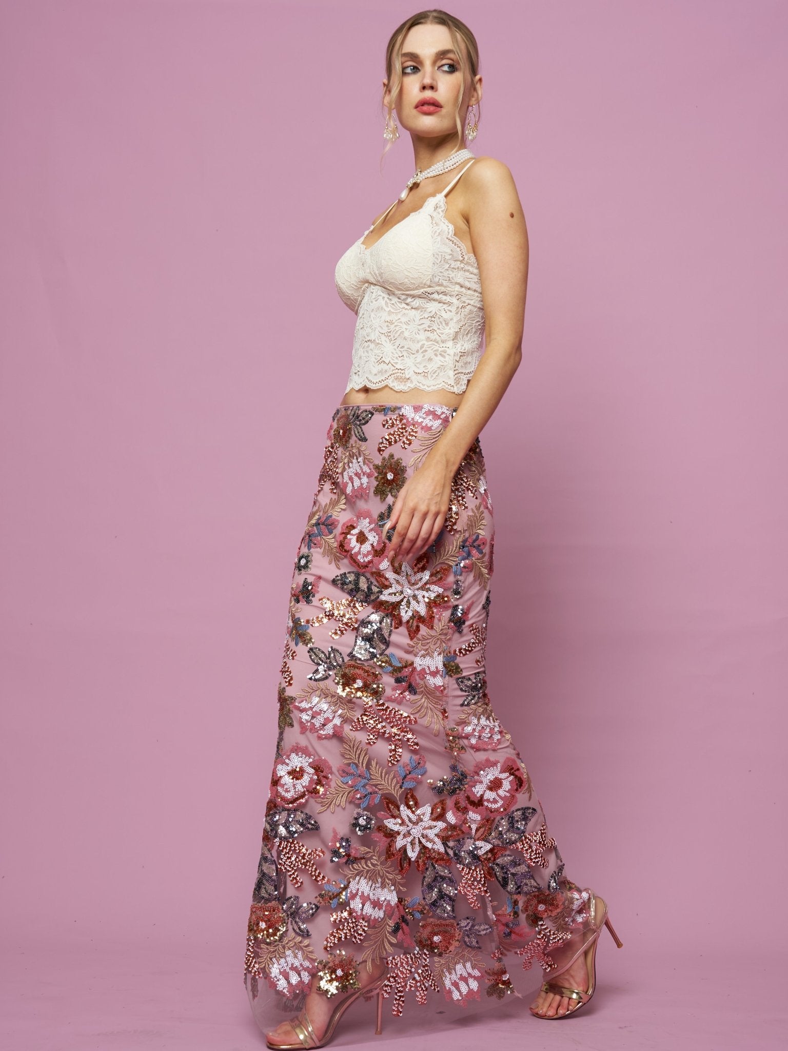 A woman stands against a pink background wearing a white lace camisole, pink heels, and the Social Queen Floral Embroidered Sequin Mermaid Skirt.