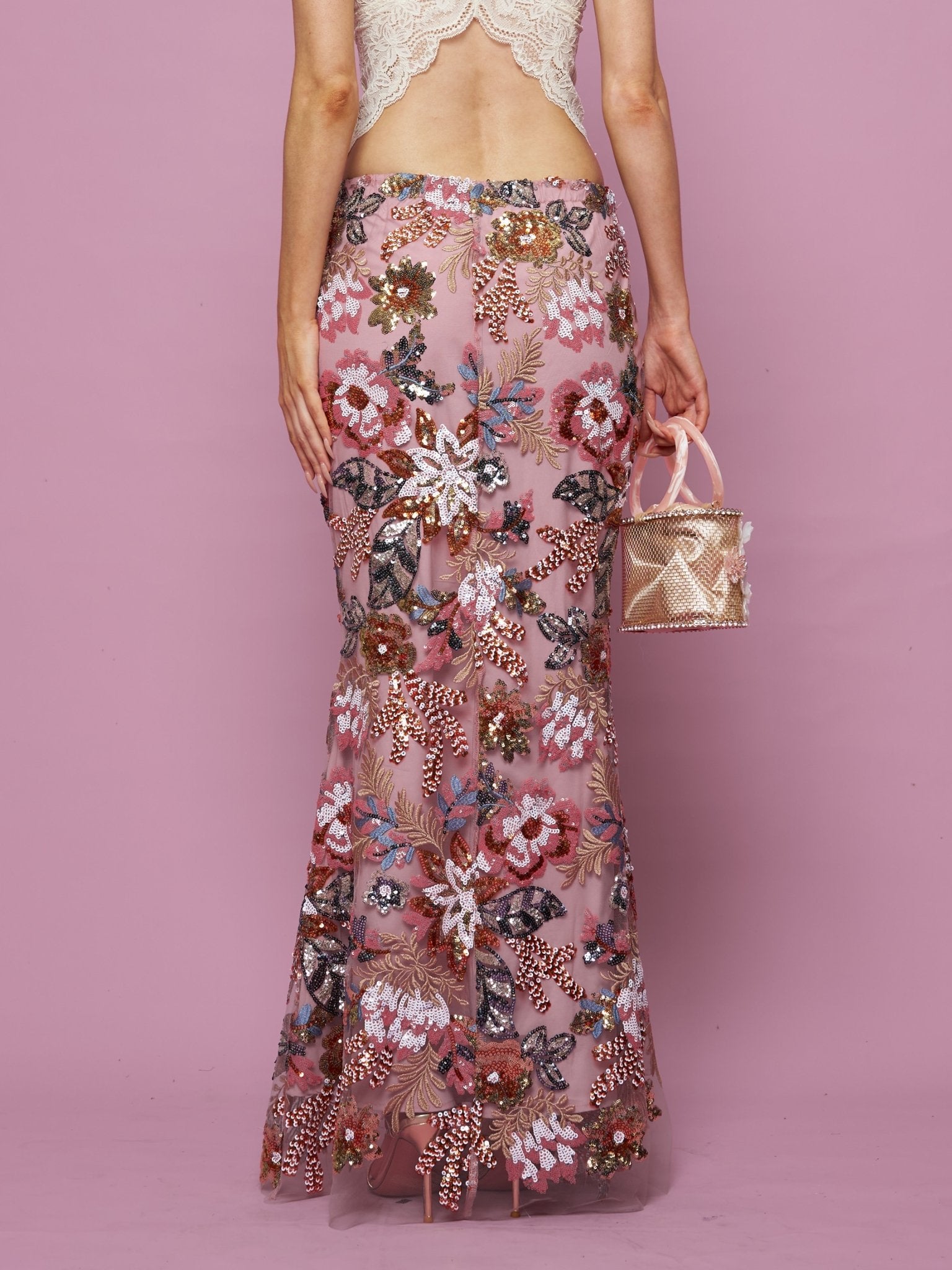 Against a pink background, a person wears the Social Queen Floral Embroidered Sequin Mermaid Skirt with a backless lace top and holds a small pink handbag.