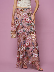 A woman stands against a pink background wearing the Social Queen Floral Embroidered Sequin Mermaid Skirt, paired with a cream lace top and holding a small pink purse.
