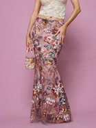 A person stands against a pink background wearing Social Queen's Floral Embroidered Sequin Mermaid Skirt, paired with a cream lace top and holding a small matching handbag.