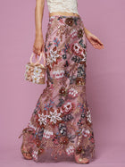 A person stands against a pink background wearing Social Queen's Floral Embroidered Sequin Mermaid Skirt, a white lace crop top, and holding a small beaded handbag.