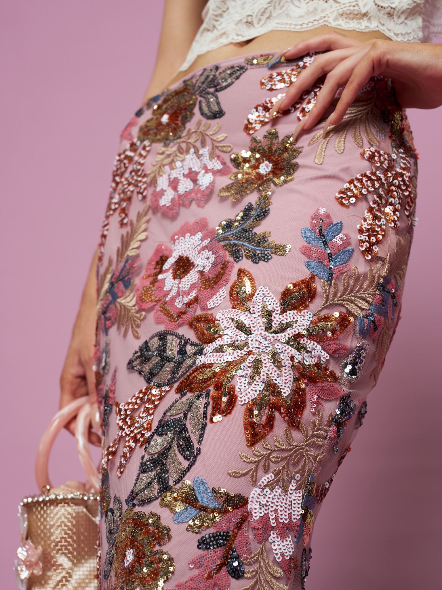 A person wears the Social Queen Floral Embroidered Sequin Mermaid Skirt and holds a small beaded handbag against a pink background.