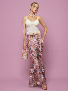 A woman poses against a pink background wearing the Social Queen Floral Embroidered Sequin Mermaid Skirt, paired with a white lace top, hoop earrings, sandals, and carrying a small pink handbag.
