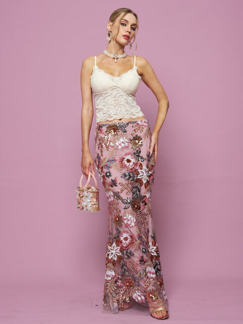 A woman poses against a pink background wearing the Social Queen Floral Embroidered Sequin Mermaid Skirt, paired with a white lace top, hoop earrings, sandals, and carrying a small pink handbag.