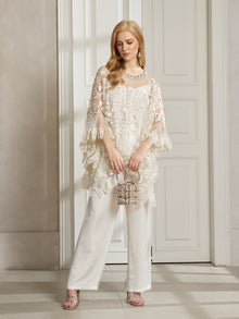 A woman stands in a well-lit room by a white door, wearing the Social Queen Floral Embroidered Sheer Overlay Top over white pants and holding a beaded handbag.