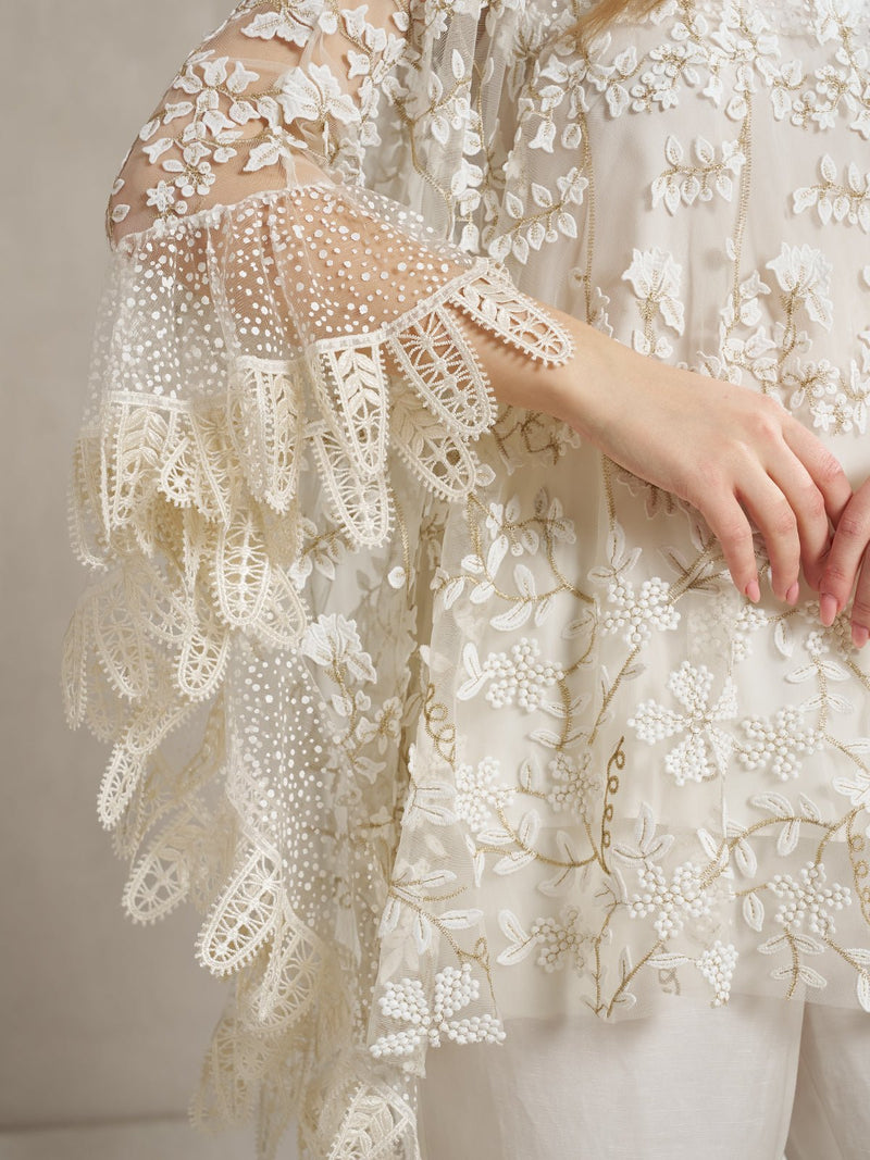A person wears the Social Queen Floral Embroidered Sheer Overlay Top, featuring delicate lace, beadwork on sleeves and bodice, and an elegant cape silhouette.