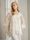 A woman with long blonde hair wears the Social Queen Floral Embroidered Sheer Overlay Top with wide sleeves, paired with white pants, standing indoors by a white-paneled wall.