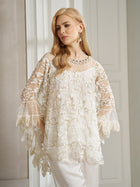 A woman with long blonde hair wears the Social Queen Floral Embroidered Sheer Overlay Top with wide sleeves, paired with white pants, standing indoors by a white-paneled wall.