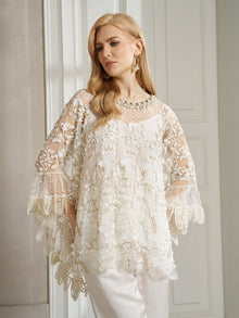 A woman with long blonde hair wears the Social Queen Floral Embroidered Sheer Overlay Top with wide sleeves, paired with white pants, standing indoors by a white-paneled wall.