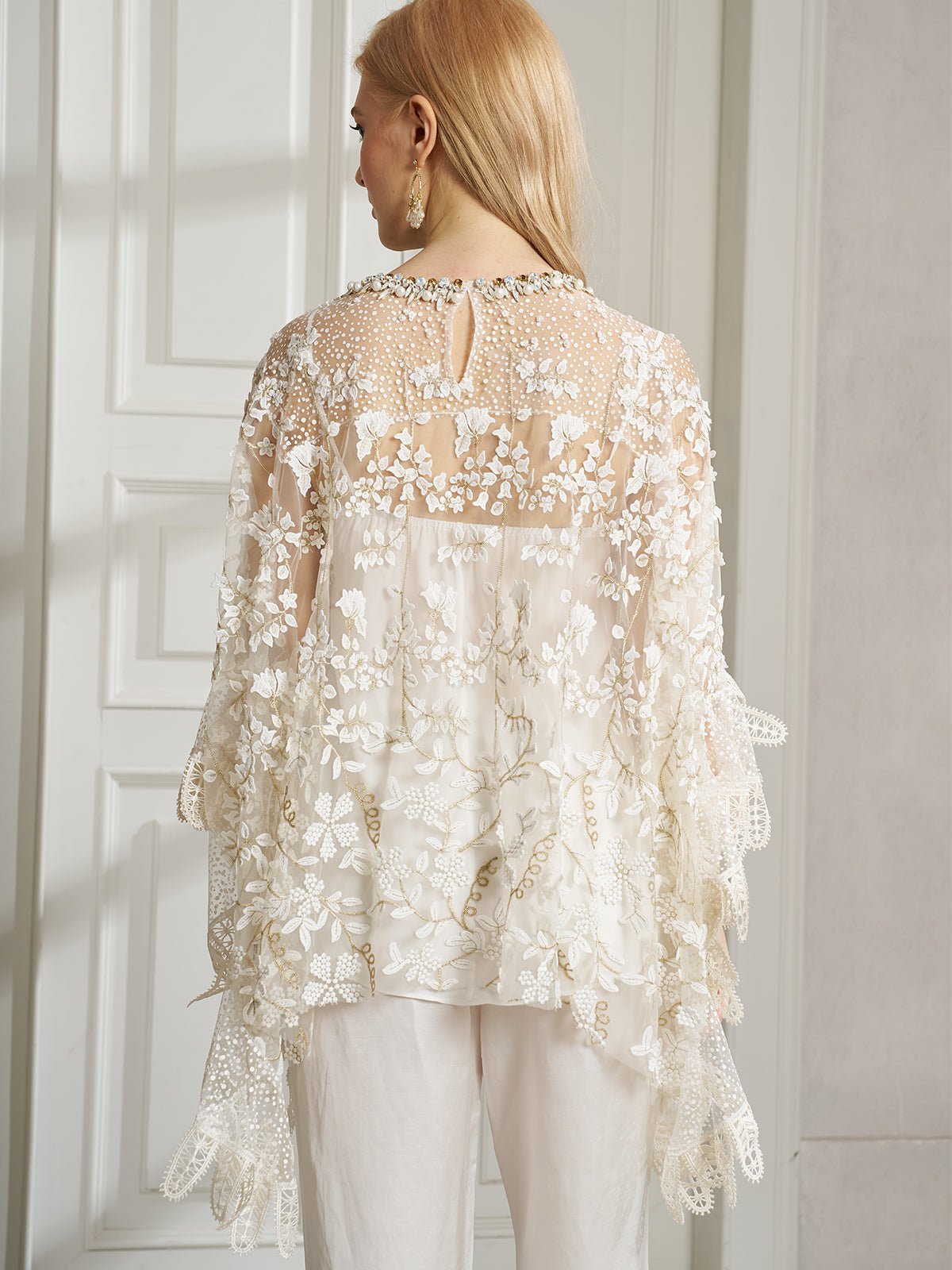 A woman, seen from behind indoors, wears the Social Queen Floral Embroidered Sheer Overlay Top paired with crisp white pants.