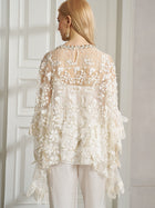 A woman, seen from behind indoors, wears the Social Queen Floral Embroidered Sheer Overlay Top paired with crisp white pants.