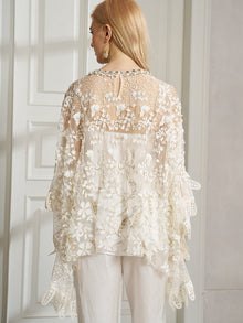 A woman, seen from behind indoors, wears the Social Queen Floral Embroidered Sheer Overlay Top paired with crisp white pants.