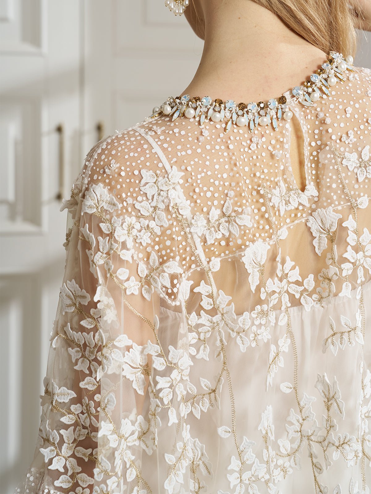 A person wears the Social Queen Floral Embroidered Sheer Overlay Top, paired with a pearl and gemstone necklace, standing indoors near white paneled doors.