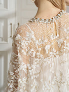 A person wears the Social Queen Floral Embroidered Sheer Overlay Top, paired with a pearl and gemstone necklace, standing indoors near white paneled doors.