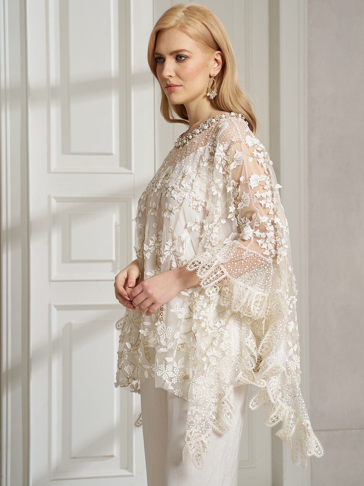 A woman stands indoors, slightly turned to the side, wearing the Social Queen Floral Embroidered Sheer Overlay Top with a cape silhouette over white pants.