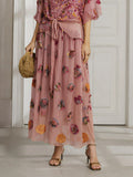 A person wears the Social Queen Floral Embroidered Silk A-Line Skirt, holding a woven bag and standing in sandals before a white door.