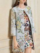 A woman in the Social Queen Floral Embroidered Silk Jacquard Jacket over a white gem-embellished dress holds a matching clutch, standing against a beige wall.