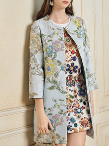 A woman in the Social Queen Floral Embroidered Silk Jacquard Jacket over a white gem-embellished dress holds a matching clutch, standing against a beige wall.