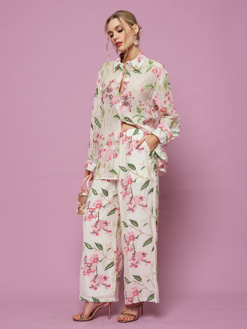 A woman stands against a pink background wearing Social Queen's Floral Embroidered Textured Wide-Leg Pants with a loose blouse, holding a small purse and wearing high-heeled sandals.