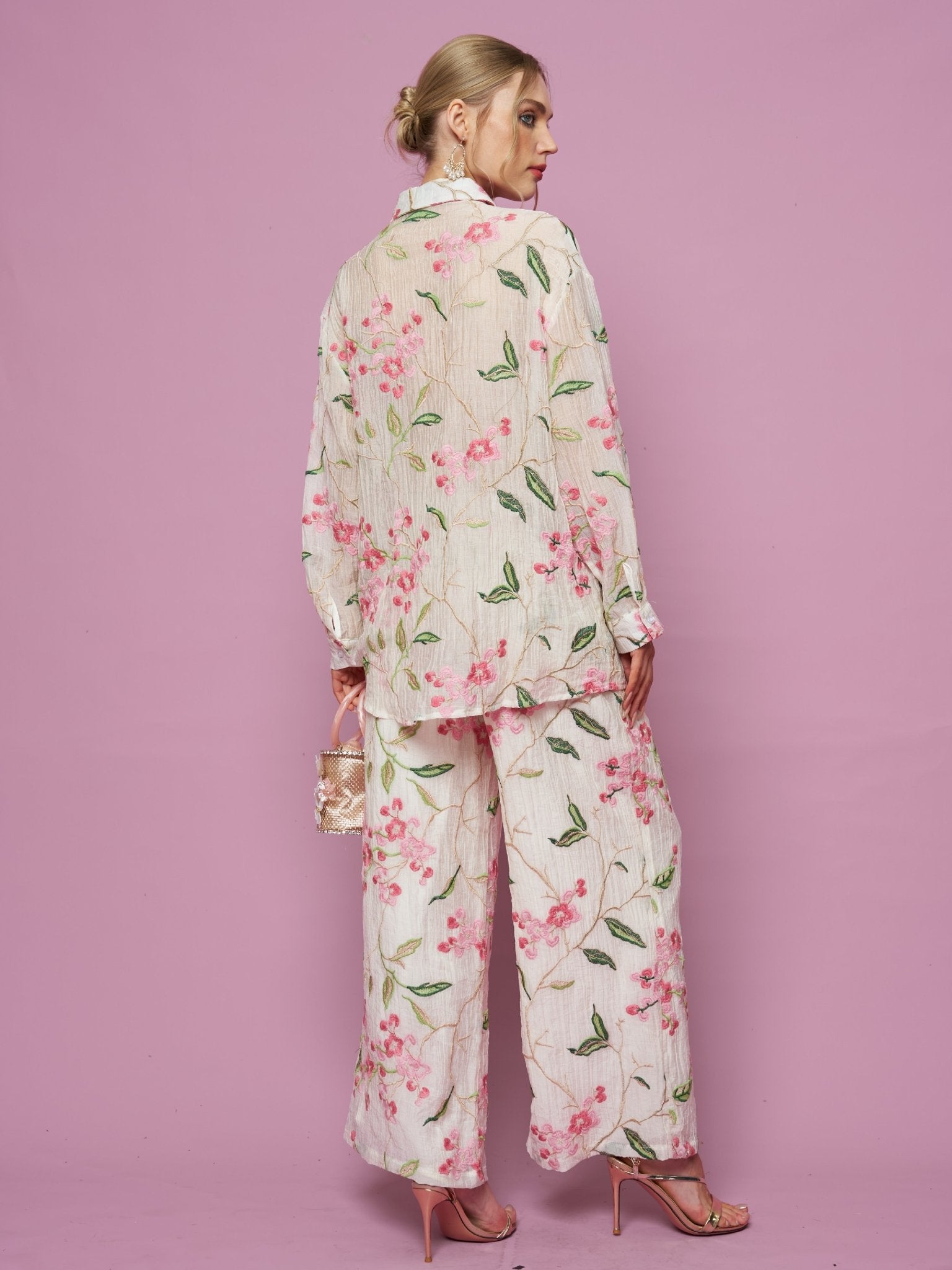 A woman stands against a pink background, wearing Social Queen's Floral Embroidered Textured Wide-Leg Pants, styled with a loose top, small transparent handbag, and pink high-heeled sandals.