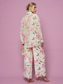 A woman stands against a pink background, wearing Social Queen's Floral Embroidered Textured Wide-Leg Pants, styled with a loose top, small transparent handbag, and pink high-heeled sandals.