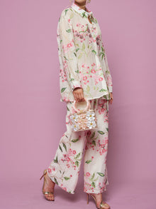 A person stands against a pink background in gold heeled sandals, wearing Social Queen’s Floral Embroidered Textured Wide-Leg Pants, a loose Tencel floral-patterned top, and holding a small decorative handbag.