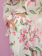 A person wearing Social Queen’s Floral Embroidered Textured Wide-Leg Pants and a matching cream outfit with pink and green details stands with one hand in their pocket against a pink background.