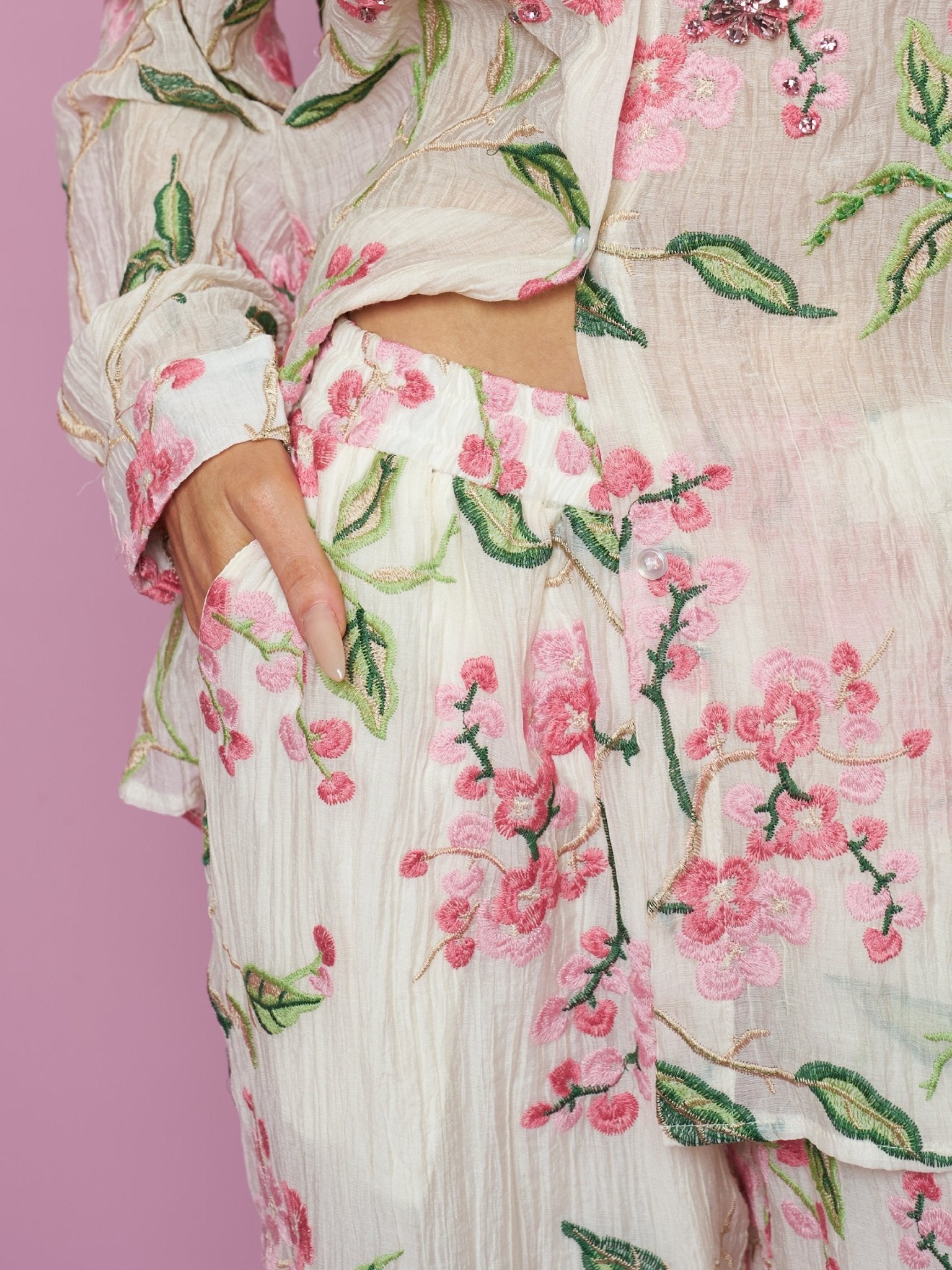A person wearing Social Queen’s Floral Embroidered Textured Wide-Leg Pants and a matching cream outfit with pink and green details stands with one hand in their pocket against a pink background.