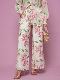 A person poses against a pink background wearing Social Queen’s Floral Embroidered Textured Wide-Leg Pants with a matching top, styled with a small bag and gold high-heeled sandals.